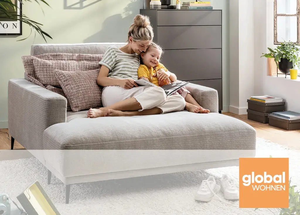 Mutter und Kind sitzen entspannt auf einer hellen Wohnlandschaft und lesen gemeinsam ein Buch. Moderne Polstermöbel von global wohnen schaffen Komfort und Familienfreundlichkeit im Wohnzimmer.